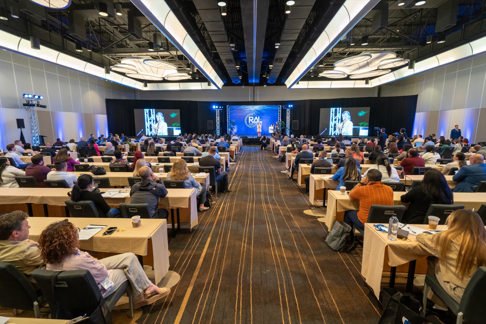 IMAG screens flanking a conference stage, where a same-day edit is played back to the room