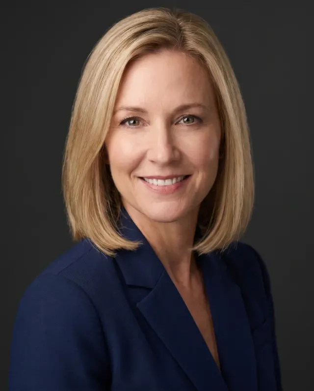 Executive headshot of a woman with a blonde bob in a navy blazer on a charcoal backdrop