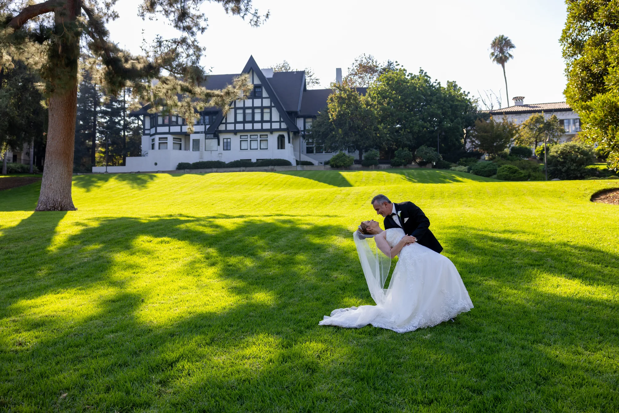 Wide view of the couple on the lawn before the half-timbered house at Ambassador Gardens, Pasadena